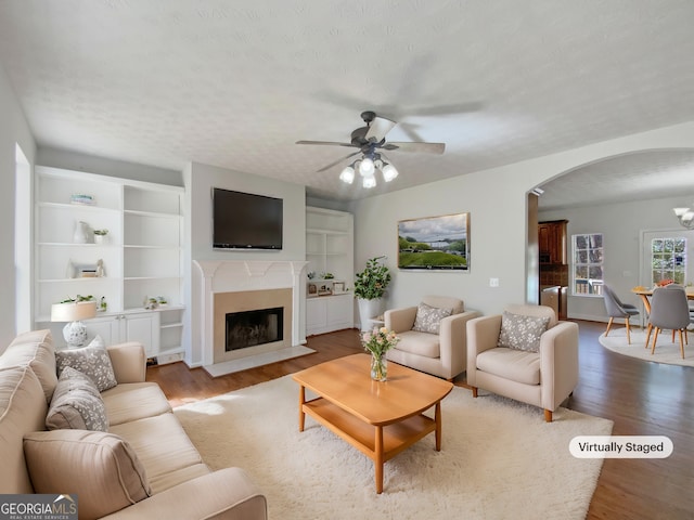 living area with a ceiling fan, wood finished floors, a fireplace with flush hearth, arched walkways, and a textured ceiling