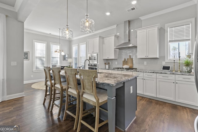 kitchen with visible vents, a center island with sink, ornamental molding, appliances with stainless steel finishes, and wall chimney exhaust hood