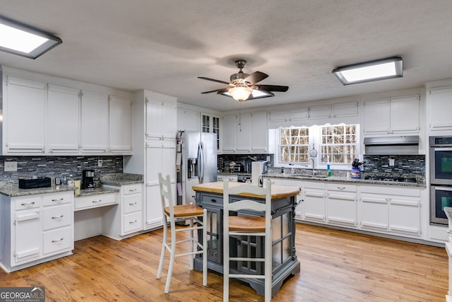 kitchen with light wood finished floors, decorative backsplash, white cabinets, under cabinet range hood, and appliances with stainless steel finishes