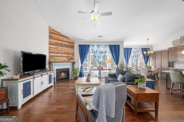 living room with visible vents, ceiling fan with notable chandelier, a warm lit fireplace, dark wood-style flooring, and vaulted ceiling