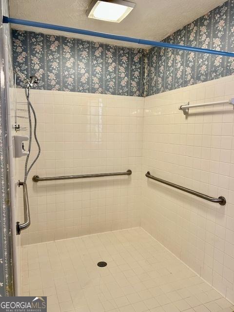 full bathroom with tiled shower, a textured ceiling, wainscoting, and wallpapered walls