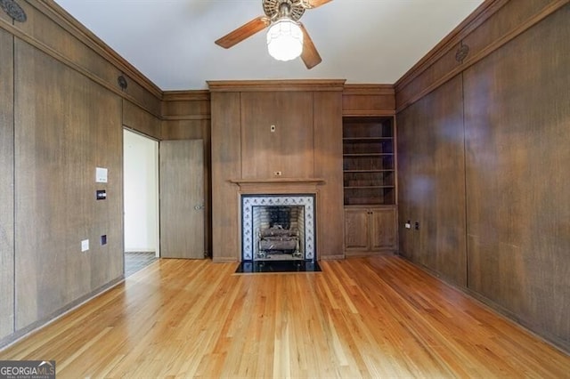 unfurnished living room with a fireplace with flush hearth, built in shelves, wooden walls, light wood finished floors, and ceiling fan