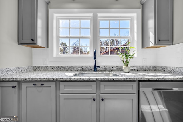 kitchen featuring tasteful backsplash, gray cabinetry, light stone countertops, dishwasher, and a sink