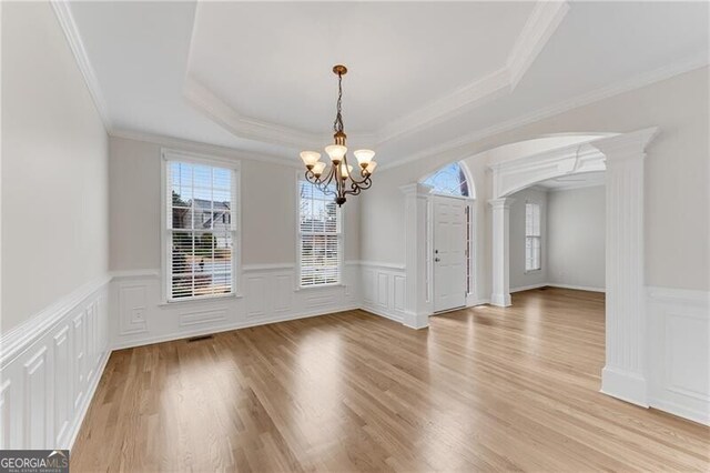unfurnished dining area with light wood-type flooring, arched walkways, an inviting chandelier, decorative columns, and a raised ceiling