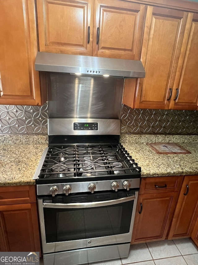 kitchen with tasteful backsplash, light stone countertops, gas range, brown cabinets, and range hood