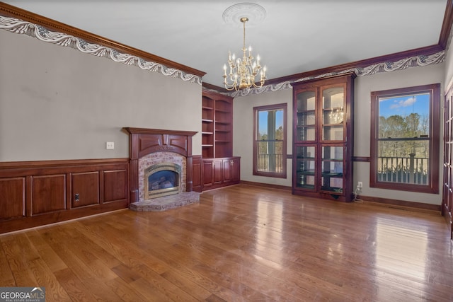 unfurnished living room with a brick fireplace, a wainscoted wall, ornamental molding, wood finished floors, and a notable chandelier