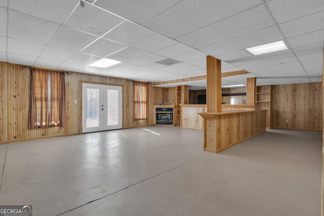 basement featuring baseboards, a drop ceiling, french doors, wood walls, and a glass covered fireplace