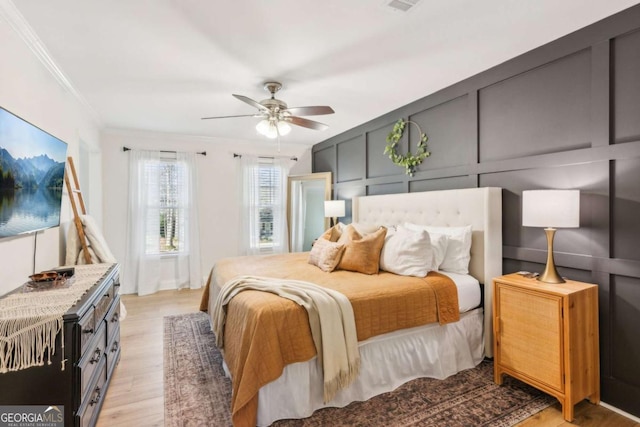 bedroom with light wood-type flooring, a ceiling fan, crown molding, and a decorative wall