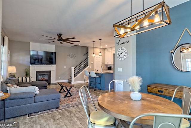 dining area featuring ceiling fan with notable chandelier, a glass covered fireplace, wood finished floors, stairway, and baseboards