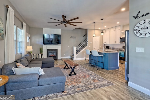 living area featuring stairway, wood finished floors, baseboards, ceiling fan, and a glass covered fireplace