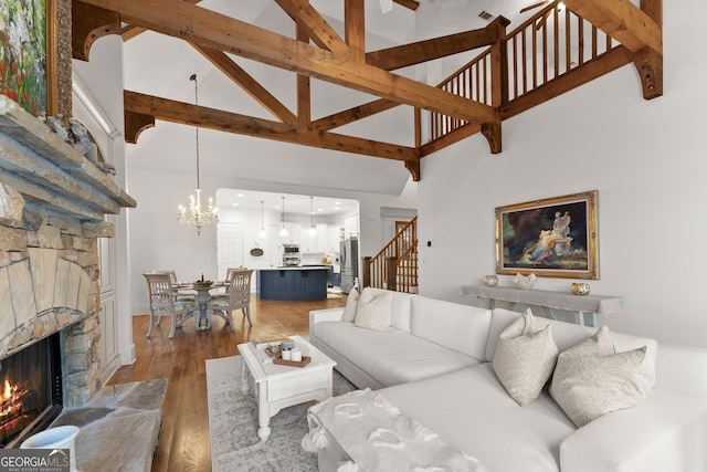 living room featuring beam ceiling, light wood-style flooring, stairway, a stone fireplace, and an inviting chandelier