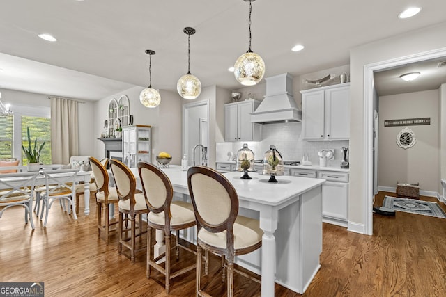 kitchen with an island with sink, wood finished floors, light countertops, decorative backsplash, and custom exhaust hood