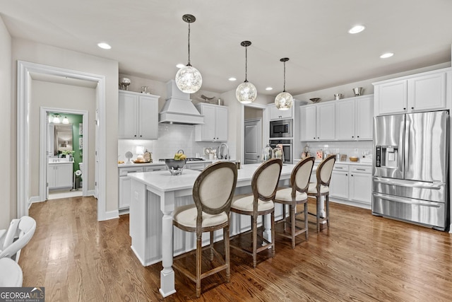 kitchen with a center island with sink, light countertops, custom range hood, appliances with stainless steel finishes, and wood finished floors