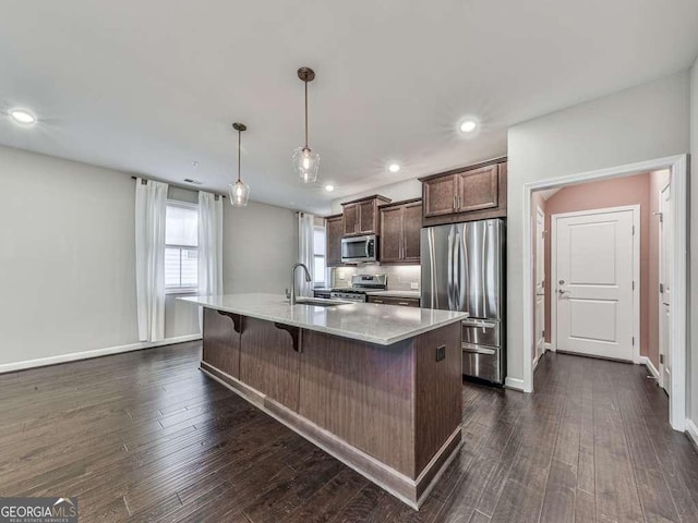 kitchen with an island with sink, dark wood-style flooring, a sink, stainless steel appliances, and a kitchen bar