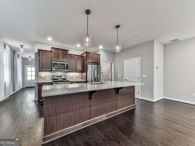 kitchen featuring dark wood-style flooring, a large island with sink, stainless steel appliances, decorative backsplash, and dark brown cabinetry