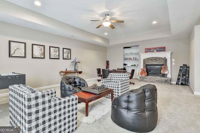 carpeted living area featuring a ceiling fan, a tray ceiling, recessed lighting, a stone fireplace, and baseboards