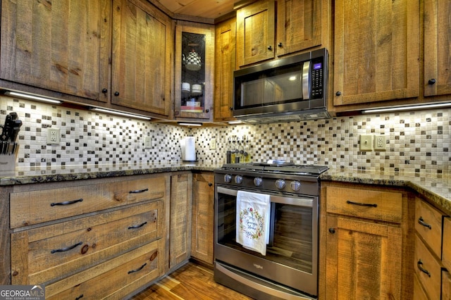 kitchen with dark stone countertops, wood finished floors, stainless steel appliances, decorative backsplash, and brown cabinets