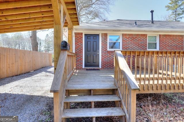 view of exterior entry with brick siding, a wooden deck, a shingled roof, and fence