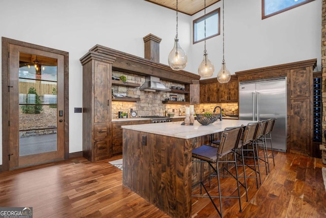 kitchen featuring dark wood-style floors, open shelves, pendant lighting, wall chimney range hood, and stainless steel built in refrigerator