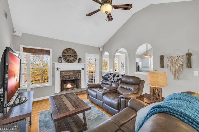 living area featuring a ceiling fan, baseboards, high vaulted ceiling, a fireplace, and light wood-style floors