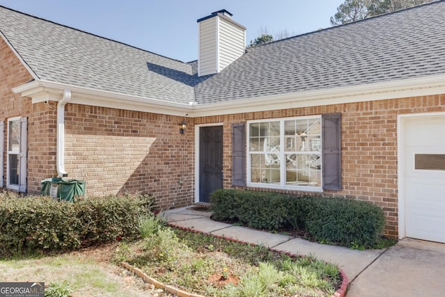 doorway to property featuring brick siding, a chimney, an attached garage, and a shingled roof