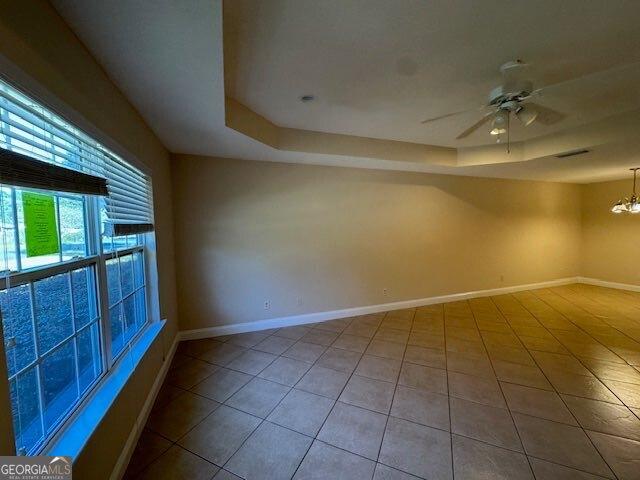 tiled empty room featuring a tray ceiling, ceiling fan with notable chandelier, and baseboards