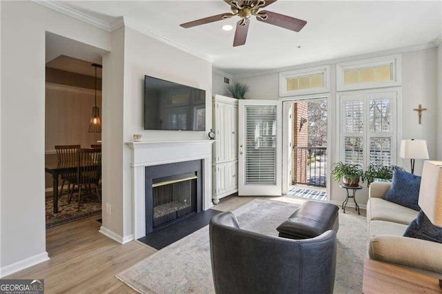 living room with a fireplace with flush hearth, ornamental molding, wood finished floors, baseboards, and ceiling fan