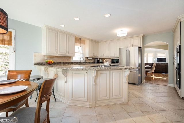 kitchen featuring stainless steel appliances, a peninsula, light tile patterned flooring, decorative backsplash, and light stone countertops