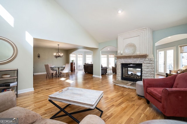 living area featuring baseboards, a stone fireplace, light wood-style flooring, a notable chandelier, and high vaulted ceiling