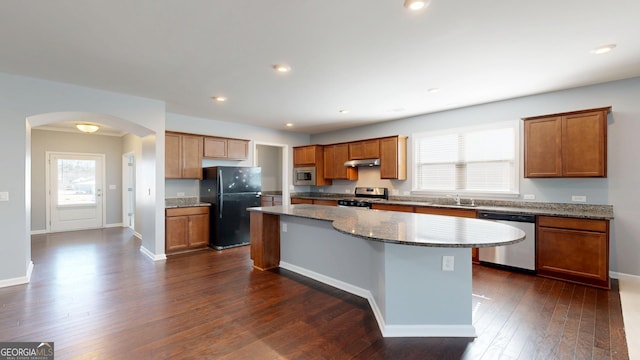 kitchen with a kitchen island, brown cabinetry, appliances with stainless steel finishes, arched walkways, and dark wood-style flooring