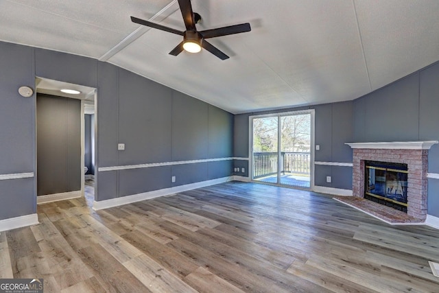 unfurnished living room with wood finished floors, a decorative wall, a brick fireplace, ceiling fan, and vaulted ceiling