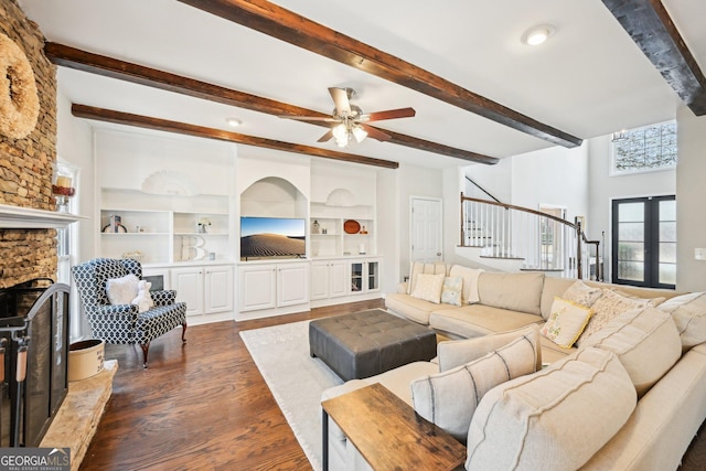 living area with beam ceiling, dark wood-style floors, a stone fireplace, ceiling fan, and stairs