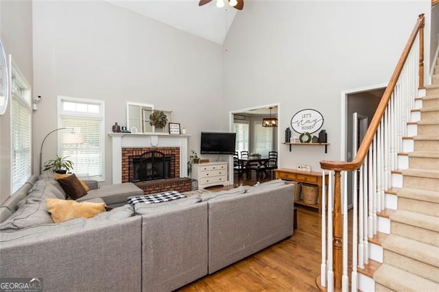 living area featuring light wood finished floors, high vaulted ceiling, ceiling fan, stairs, and a brick fireplace