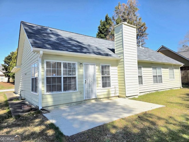 back of house with a shingled roof, a patio, a lawn, and a chimney