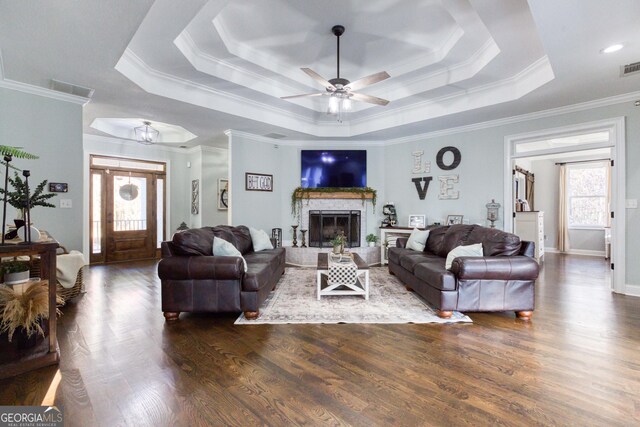 living room featuring visible vents, ornamental molding, a tray ceiling, wood finished floors, and a stone fireplace