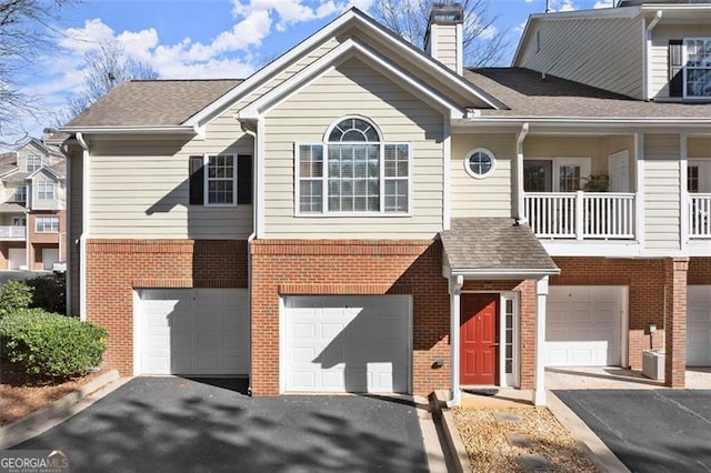 multi unit property featuring brick siding, roof with shingles, a chimney, a garage, and driveway