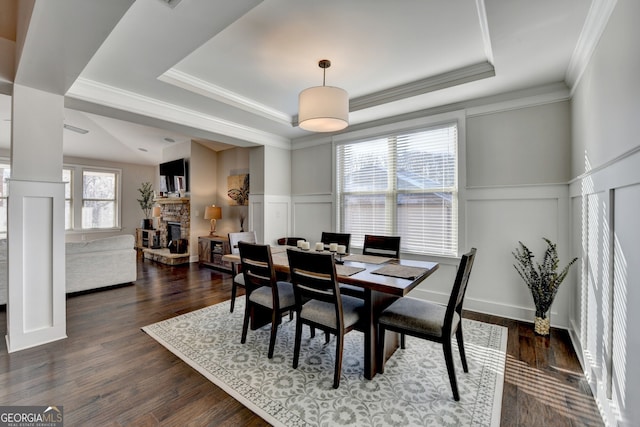 dining area featuring a wainscoted wall, a decorative wall, a stone fireplace, dark wood finished floors, and a tray ceiling