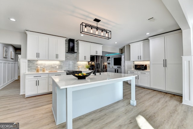 kitchen with light wood finished floors, a kitchen island with sink, decorative backsplash, wall chimney exhaust hood, and refrigerator with ice dispenser