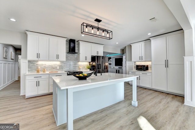 kitchen featuring light wood finished floors, wall chimney exhaust hood, tasteful backsplash, a kitchen island with sink, and fridge with ice dispenser
