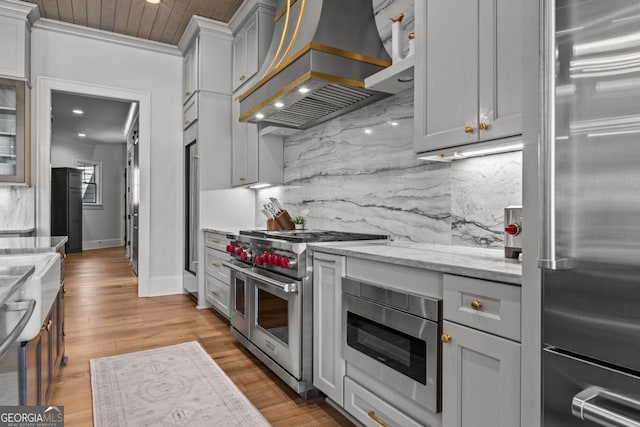 kitchen featuring light stone counters, custom exhaust hood, decorative backsplash, high quality appliances, and light wood-type flooring
