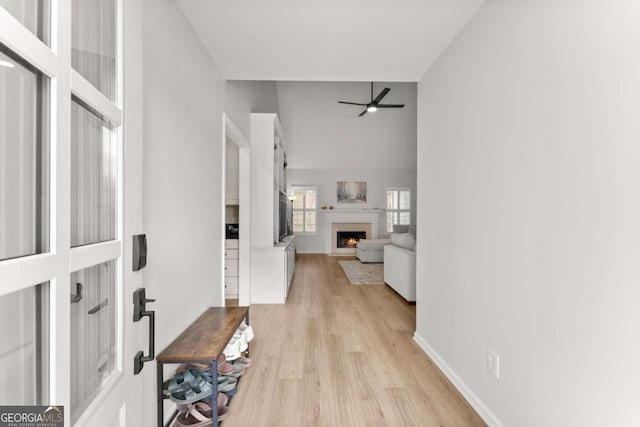 foyer entrance featuring a ceiling fan, baseboards, a high ceiling, a lit fireplace, and light wood-style floors