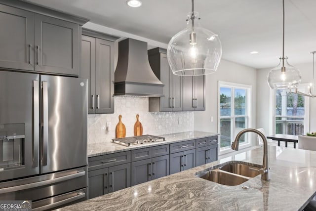 kitchen featuring gray cabinetry, a sink, stainless steel appliances, wall chimney range hood, and light stone countertops