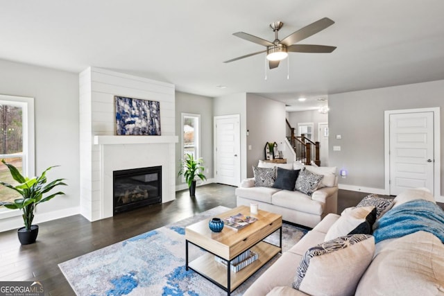 living area featuring baseboards, stairs, wood finished floors, a glass covered fireplace, and a ceiling fan