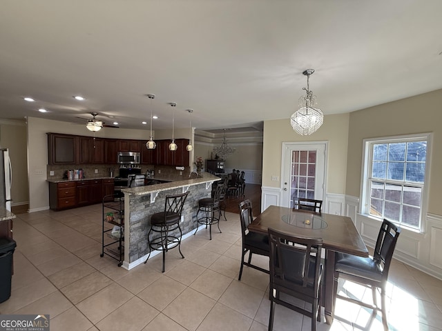 dining space with ceiling fan with notable chandelier, a wainscoted wall, recessed lighting, and light tile patterned floors