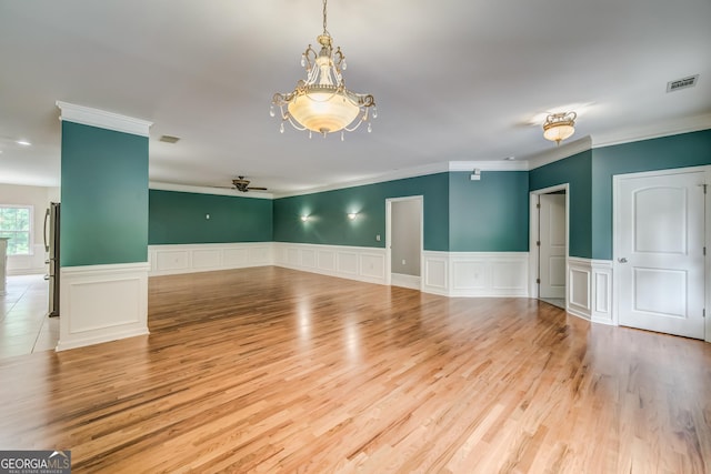 spare room featuring a wainscoted wall, wood finished floors, visible vents, ornamental molding, and ceiling fan with notable chandelier
