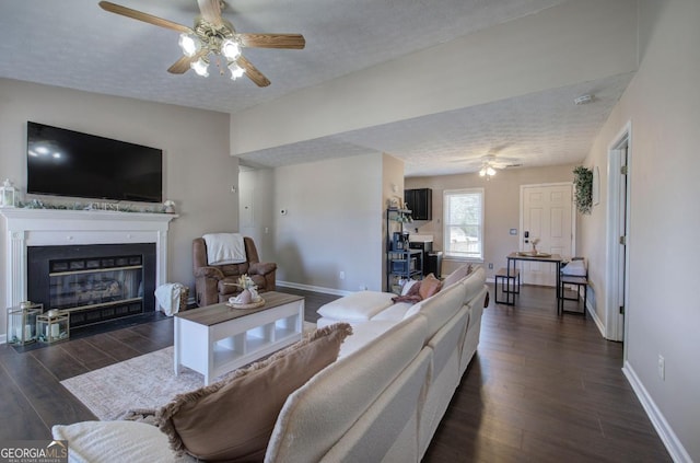 living room with a textured ceiling, a glass covered fireplace, dark wood-style floors, baseboards, and ceiling fan