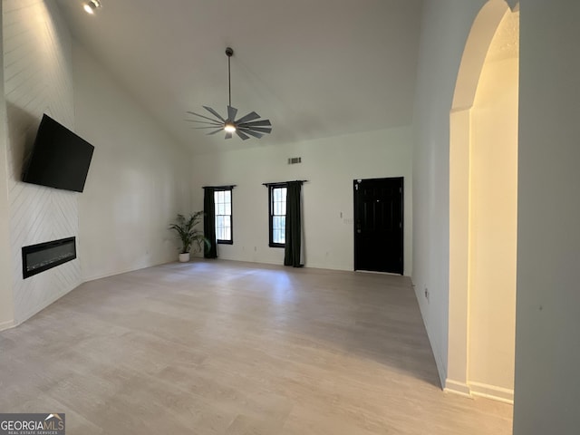 unfurnished living room featuring a ceiling fan, high vaulted ceiling, arched walkways, a glass covered fireplace, and light wood-type flooring