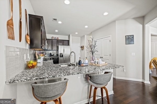 kitchen featuring dark brown cabinets, light stone countertops, stainless steel fridge with ice dispenser, decorative backsplash, and a peninsula