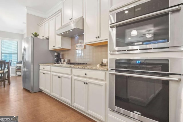 kitchen featuring light wood finished floors, stainless steel appliances, decorative backsplash, under cabinet range hood, and crown molding