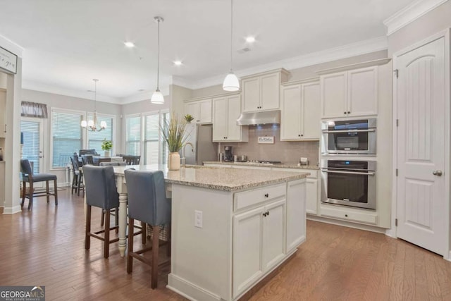 kitchen featuring wood finished floors, appliances with stainless steel finishes, under cabinet range hood, crown molding, and backsplash